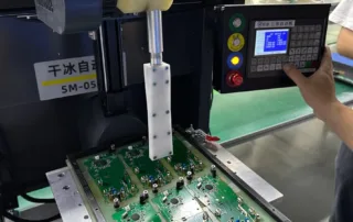 Technician operating a PCB assembly machine, pressing a green circuit board in a metal jig while adjusting the control panel display and buttons