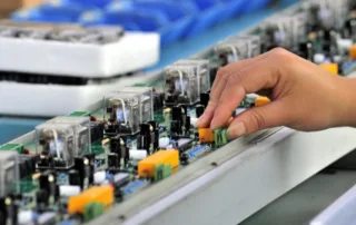 Close-up of a hand adjusting components on an exposed circuit board along a production line.