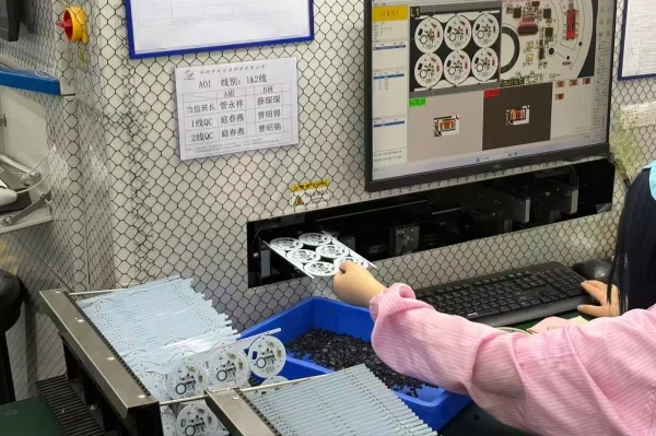 Worker places white circular components onto a blue bin in a production line, with a monitor showing diagrams in the background.