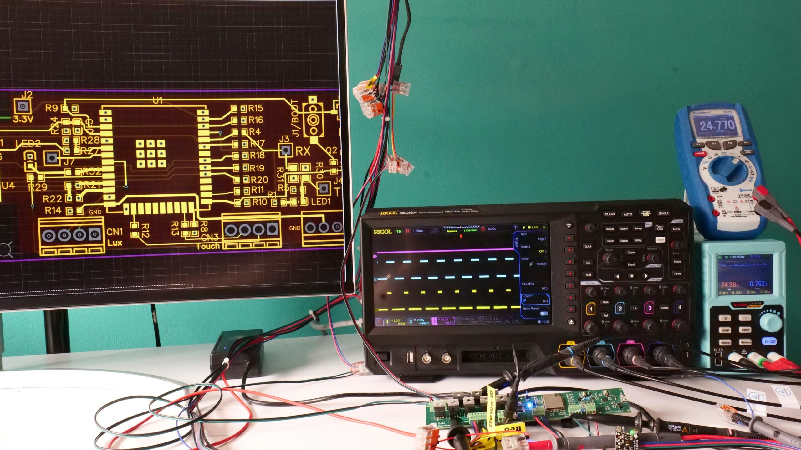 Electronics workbench with a PCB layout display, oscilloscope, multimeter, power supply and connected circuit board